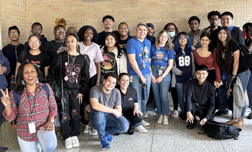 A group of students poses outside of a high school in front of a brick wall.